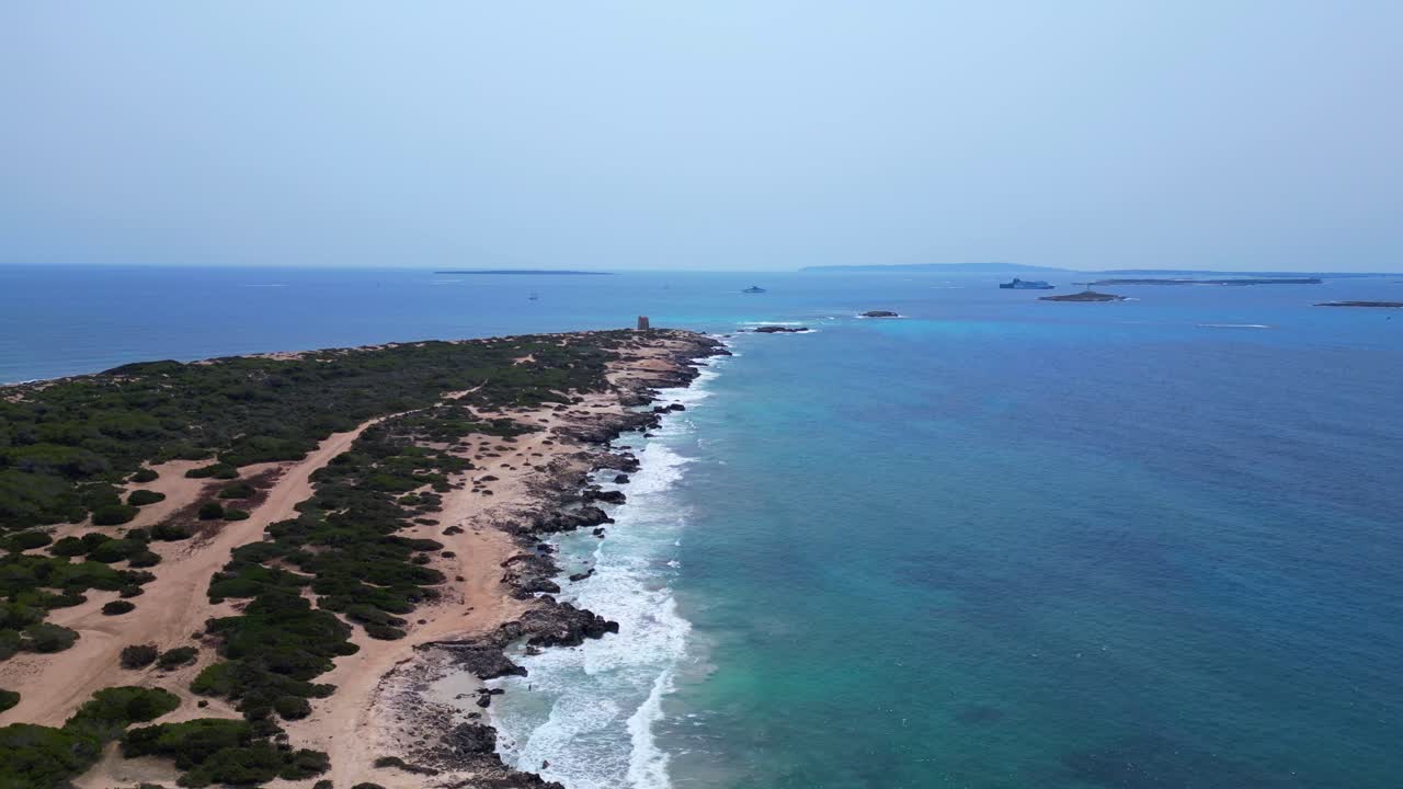 Turquoise waves crashing on the rocky coast of Ses Salines beach with the ancient watchtower. Beautiful aerial view flight descending drone