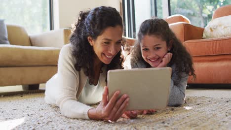 Happy-mixed-race-mother-and-daughter-laying-on-the-floor,having-fun-and-using-tablet