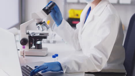 female lab research worker in ppe looking through microscope and recording results on laptop