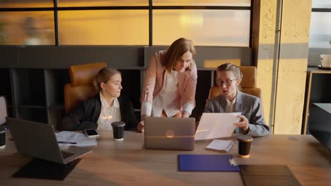 A-trio-of-confident-businesswomen-in-business-clothes-and-jackets-gathered-around-a-wooden-table-in-an-office-and-discussing-the-current-plan-and-solution-to-a-problem-using-sheets-and-a-laptop-in-a-sunny-office