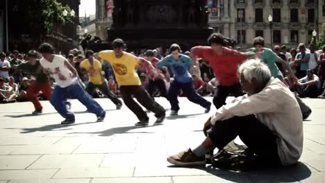 a group of young dancers showcases their energetic routine in a bustling public square. an older man sits nearby, observing the performance amid a lively audience.