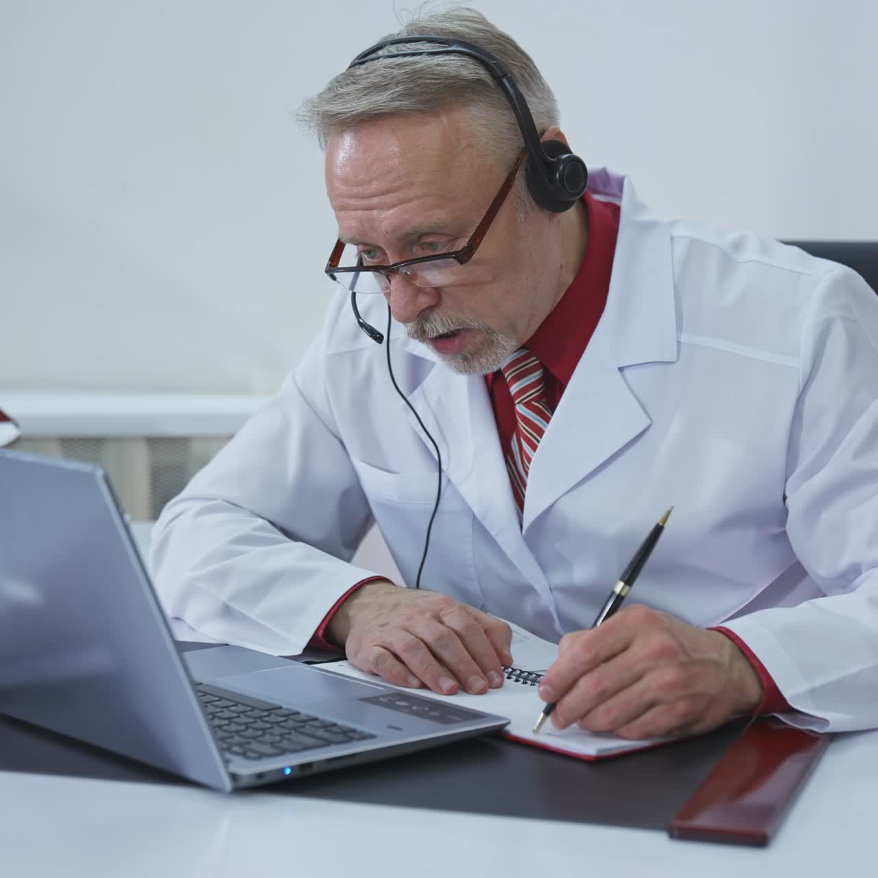 Remote medicine. Senior male specialist giving a consultation to a patient through video call. Medical specialist in headphones talking and writing treatment.