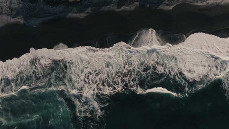 Drone top-down aerial view of ocean waves breaking onto a black sand beach alongside a rocky volcanic coastline in Iceland during winter. Coastal nature and Nordic wilderness concept
