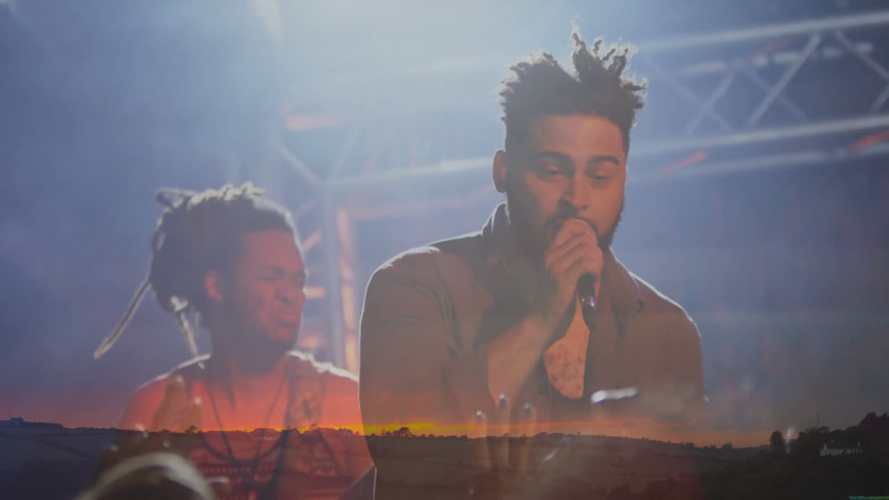 male singer and percussionist playing music on dim stage with haze, lighting rig and sunset overlay