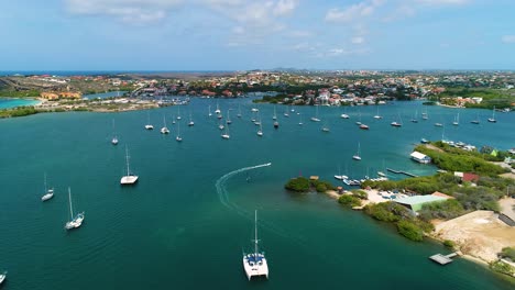 boat drives into channel of spanish waters harbor with catamaran and yachts anchored, drone rear view tracking