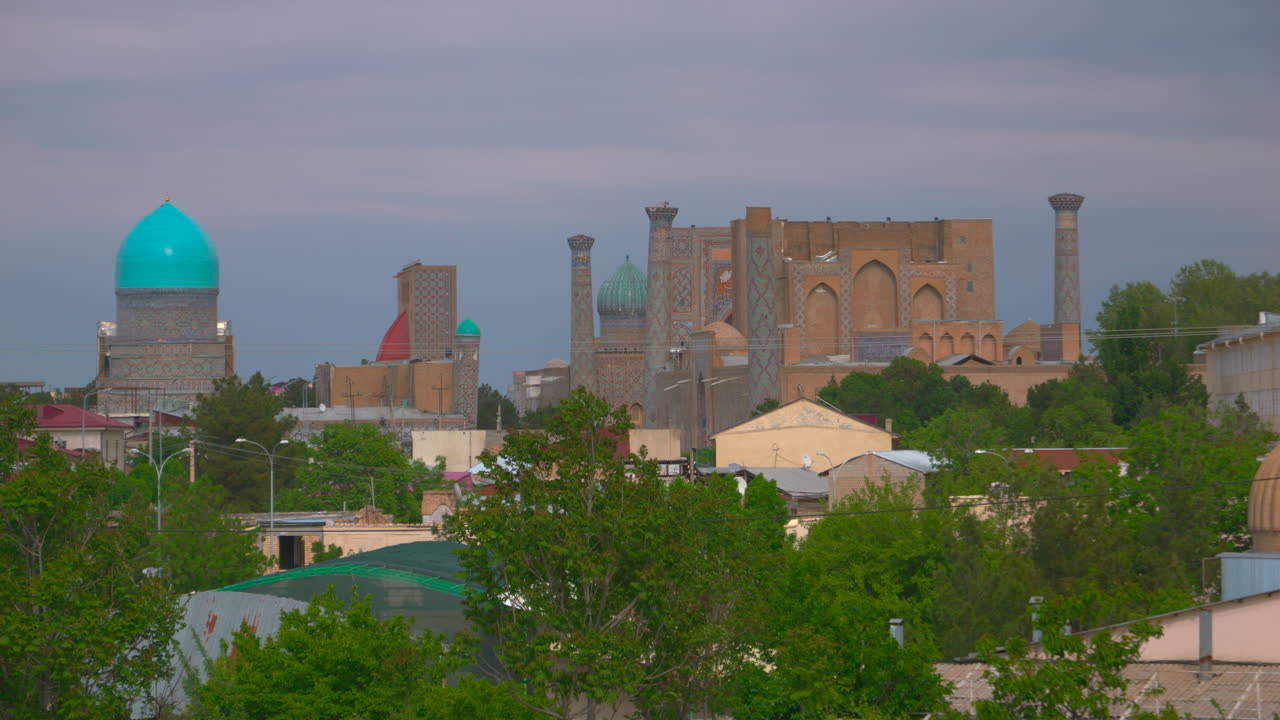 Panoramavy över en historisk stadssilhuett med turkosa kupoler, minareter och utsmyckad islamisk arkitektur under en dramatisk himmel. En centralasiatisk stadsvy med kända landmärken och gröna träd.