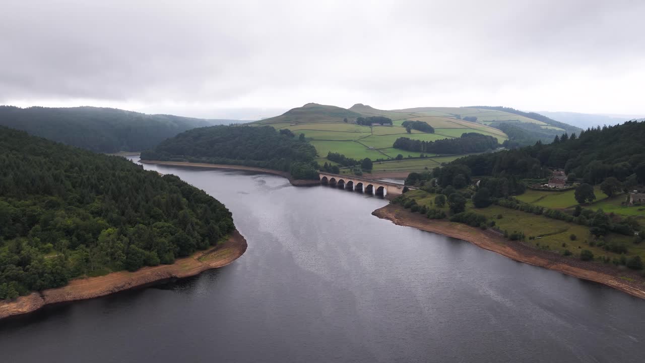 Drone glides above Ladybower Reservoir, revealing lush hills, forested banks, and a stone viaduct under overcast skies with soft, diffused daylight