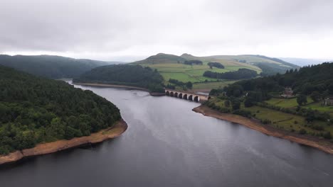 drone glides above ladybower reservoir, revealing lush hills, forested banks, and a stone viaduct under overcast skies with soft, diffused daylight