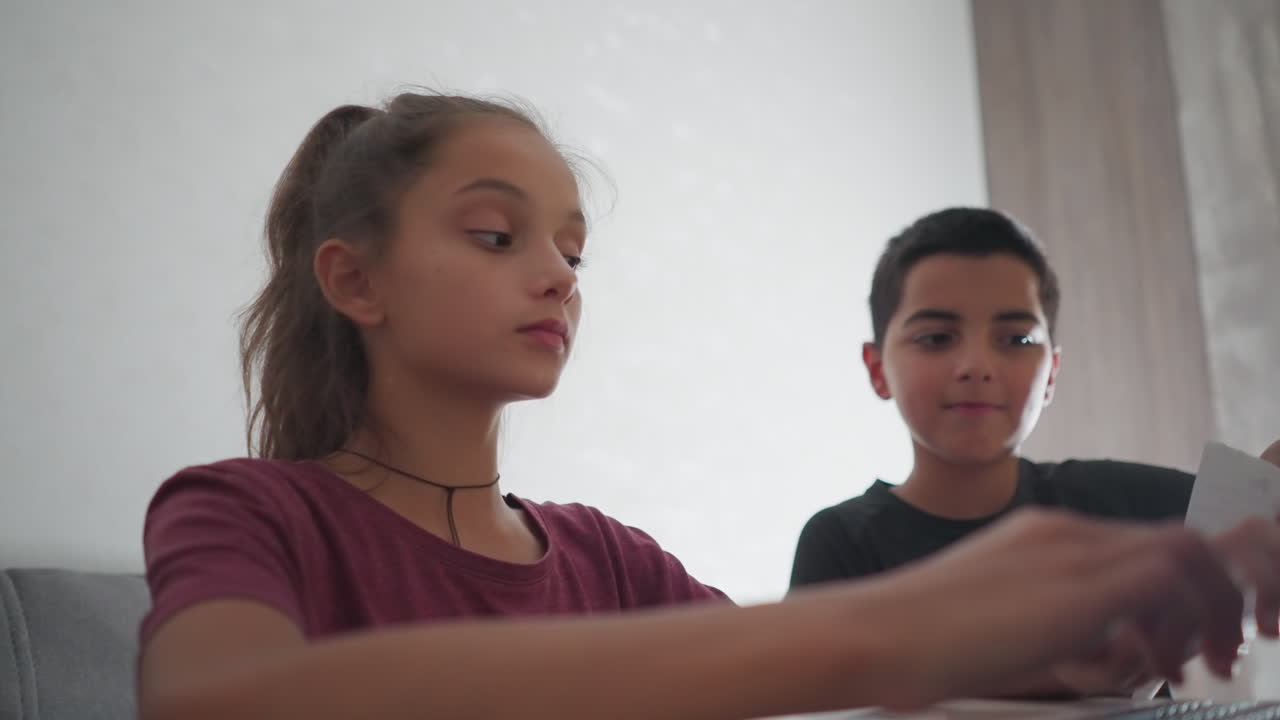 Primer plano de un niño escribiendo con un bolígrafo que toca a una niña que lleva una pulsera de cuerda roja mientras escribe en un portátil; ella se detiene para echar un vistazo a su trabajo, mostrando curiosidad e interés durante un momento de estudio compartido en casa.