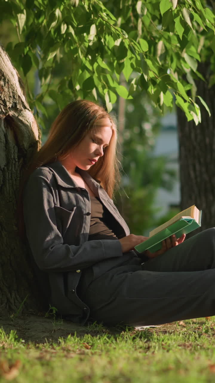 mujer sentada al aire libre con las piernas estiradas, apoyada en un árbol en un campo de hierba, leyendo un libro, las hojas de los árboles se balancean suavemente en la brisa, el fondo con vegetación y un edificio blanco