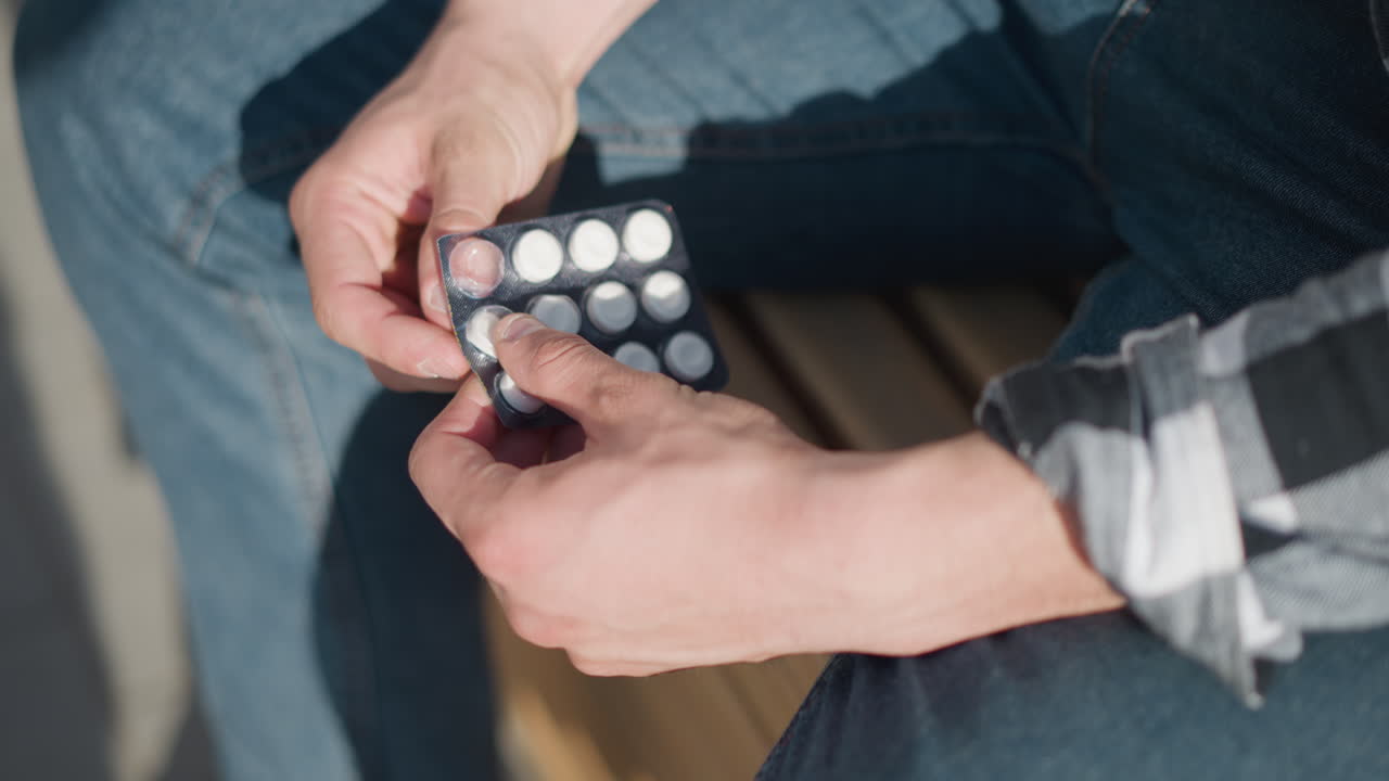 Close up of young man in plaid shirt and jeans seated on wooden bench using hands to press out tablet from blister pack under bright outdoor lighting with soft shadows on clothing and hands