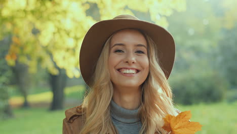 Caucasian-young-blonde-woman-wearing-a-hat-smiling-and-hiding-her-face-behind-a-yellow-autumn-leaf-in-the-park