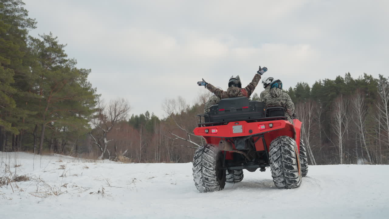 Quad rosso su sentiero innevato, due piloti caucasici da dietro con le braccia alzate in segno di trionfo, quad che attraversa una radura ghiacciata, duo con casco in giacche mimetiche, tracce di pneumatici sulla neve, ampio orizzonte di pini, euforia.