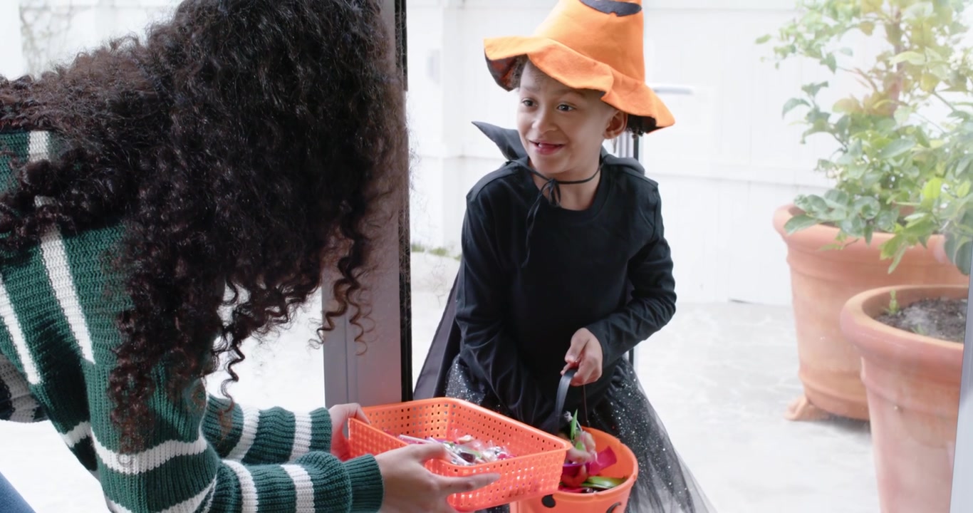 African American woman at door offering tray while child in costume filling pumpkin pail for treats
