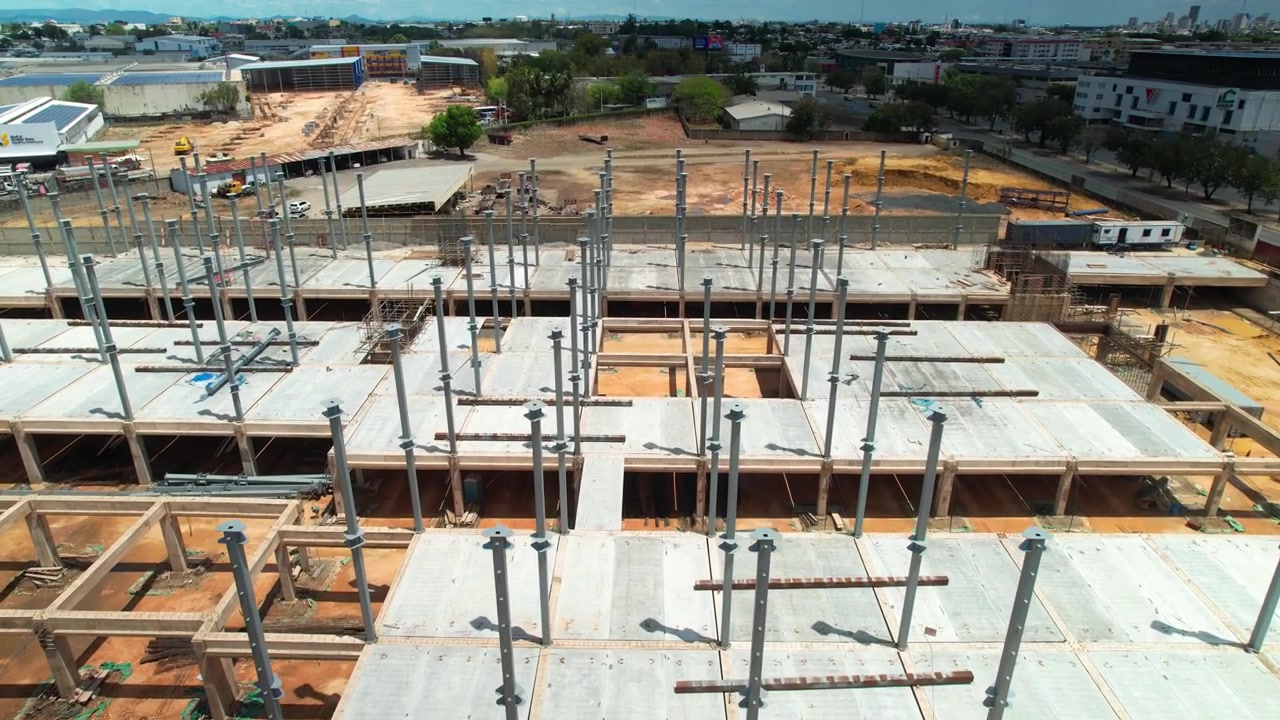 High-angle aerial drone shot flying forward and tilting down over a commercial building construction site, showcasing steel pillars, concrete floor slabs, and surroundings