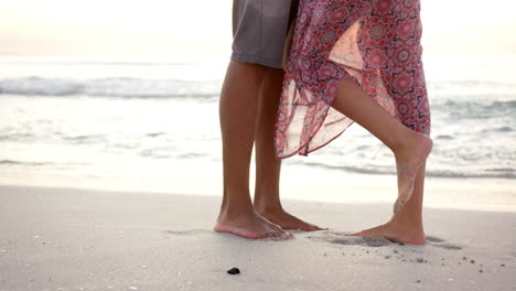 barefoot couple stands close on a sandy beach, with a woman in a patterned dress