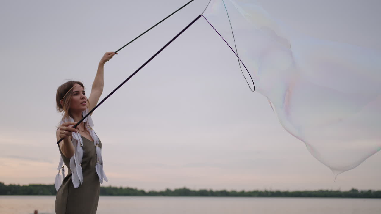 젊은 여성 예술가 는 일몰 에 호수 연안 에 거대한 비누 거품 을 불어넣는 비누 거품 쇼 를 보여준다. 느린 움직임 으로 비누 거품 의 아름다운 쇼 를 보여준다