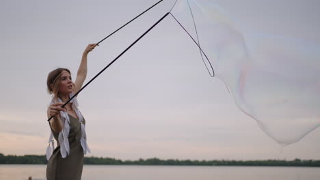 uma jovem artista mostra um espetáculo de bolhas de sabão explodindo enormes bolhas de sabão na costa de um lago ao pôr do sol. mostre um belo espetáculo de bolhas de sabão em câmera lenta