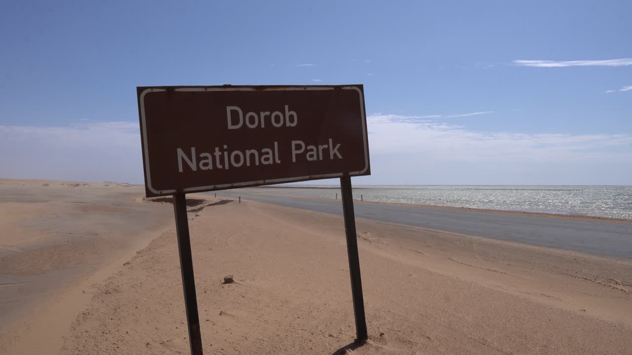signo de nombre para el parque nacional de dorob al lado de una carretera, arena soplando en el suelo en un día soleado