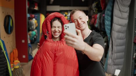 Ein-Lächelndes-Paar-Kauft-Im-Sportbekleidungsgeschäft-Ein,-Probiert-Einen-Schlafsack-Aus-Und-Macht-Ein-Selfie