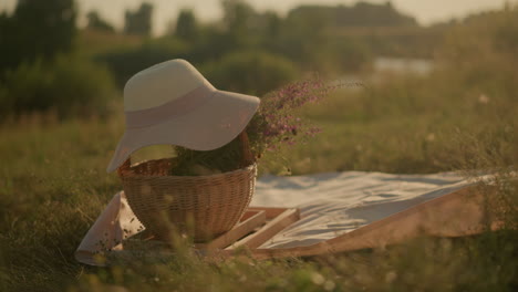 cesta tejida con flores y sombrero de sol en una alfombra en una zona de hierba abierta bajo la luz del sol, con un fondo borroso de la naturaleza, evocando un pacífico día de verano, vibraciones de picnic en el campo