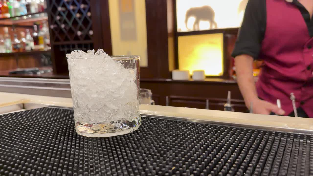 Bartender adds liquor to glass of crushed ice, warm lighting, steady close-up, upscale bar setting