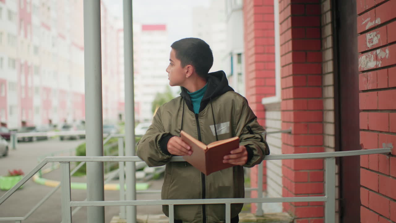 Kid in hooded jacket stands near red brick building holding brown book, resting on iron railing, looking around thoughtfully while parked cars and city buildings form background scene