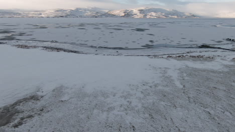 Dolly-forward-above-snowy-shore,-frozen-lake-and-mountains,-at-sunny-day,-Iceland