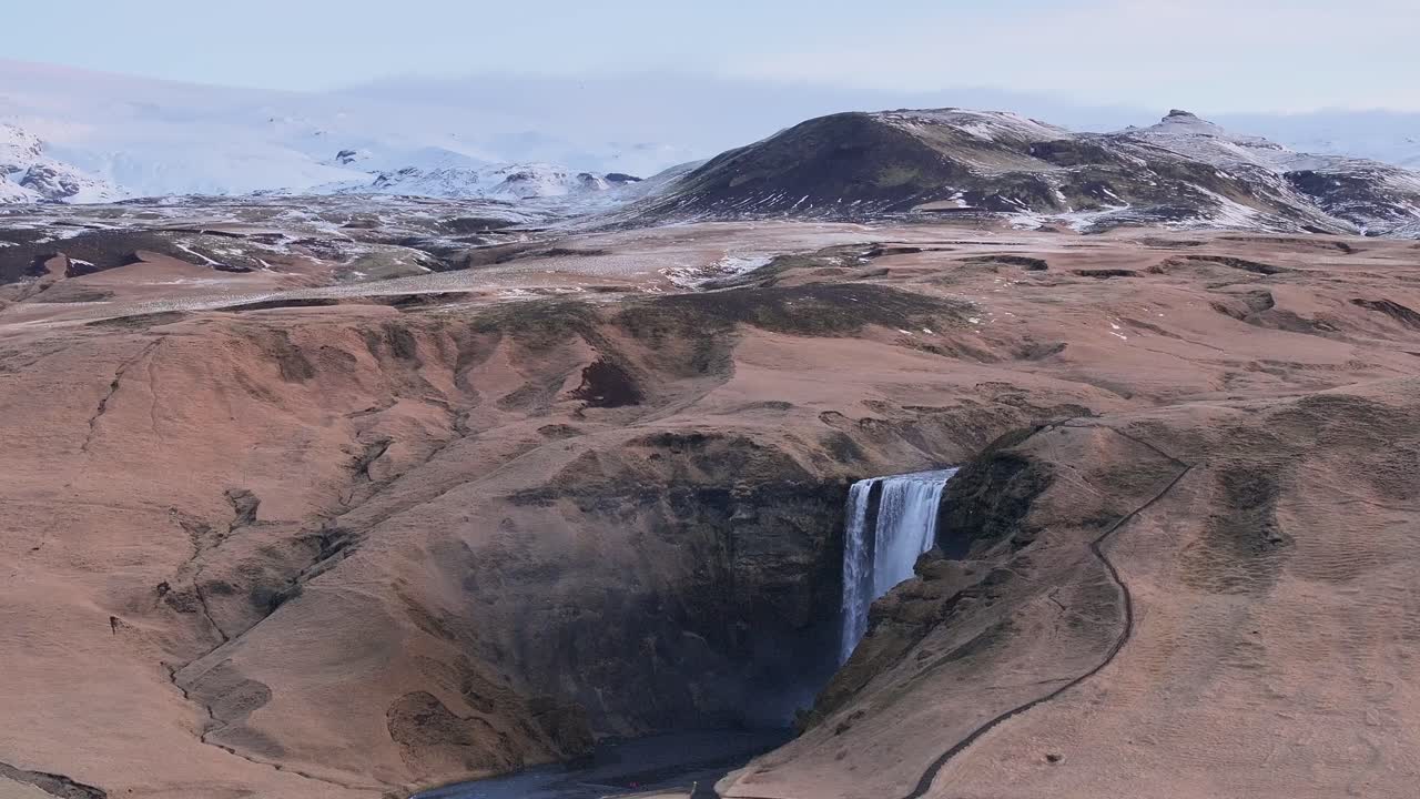 Lateral drone shot of Skógafoss waterfall in Skógar, Rangárþing eystra, Iceland, revealing the surrounding volcanic terrain and distant snowy mountains.
