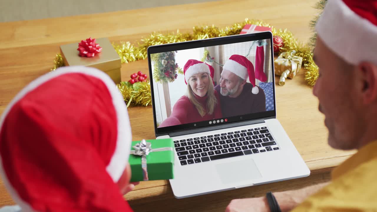 padre y hijo caucásicos con sombreros de santa usando una computadora portátil para una videollamada de navidad con una pareja en la pantalla
