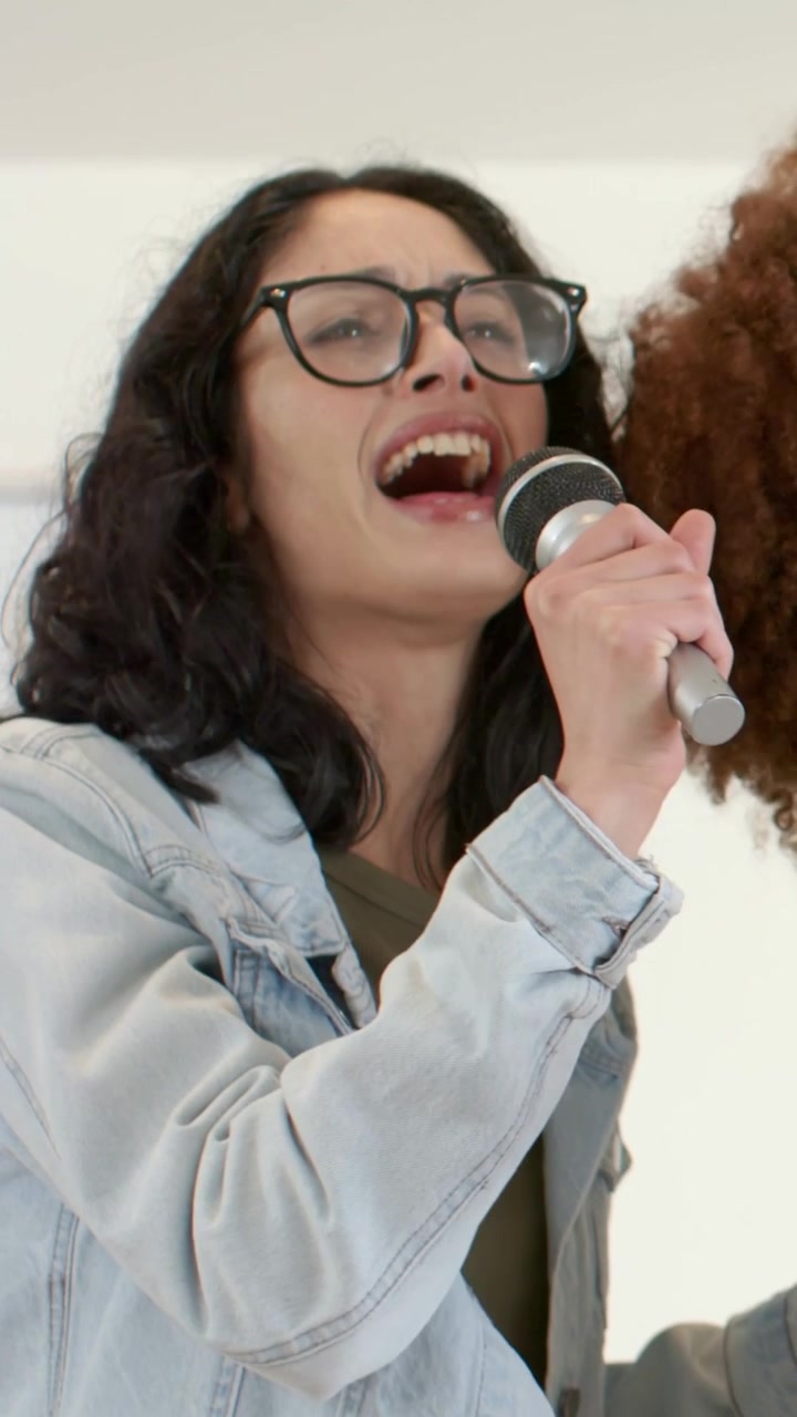 Vertical video: Song starting, 3 women rehearsing in studio, one wearing pink sweater, sharing mics