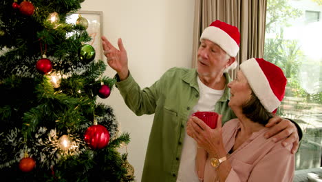 senior couple decorating christmas tree at home, wearing santa hats, smiling warmly