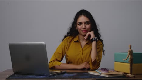 a south asian woman in yellow shirt posing confidently with hand on chin at laptop in modern office