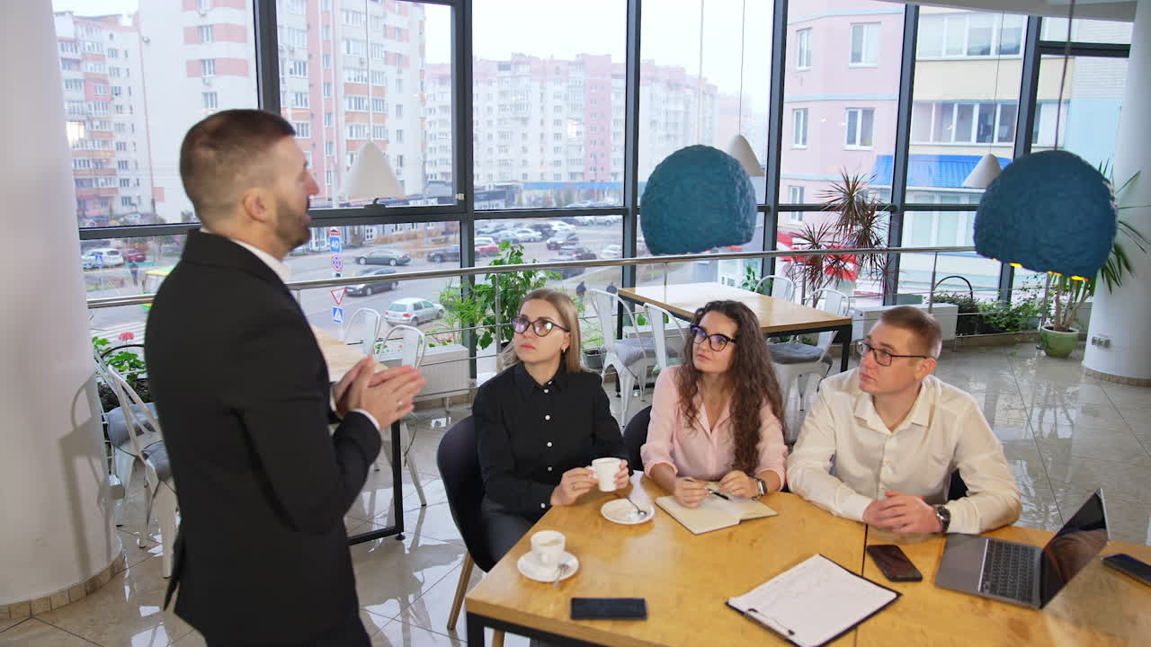 Business leader wearing black suit talks to his staff sitting in front of him. Employees listening to their boss attentively.