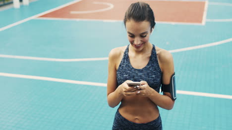 Sportliches-Mädchen-Mit-Airpods-SMS-Nachricht-Auf-Dem-Smartphone,-Während-Es-An-Einem-Sommertag-Auf-Dem-Außenplatz-Steht-1