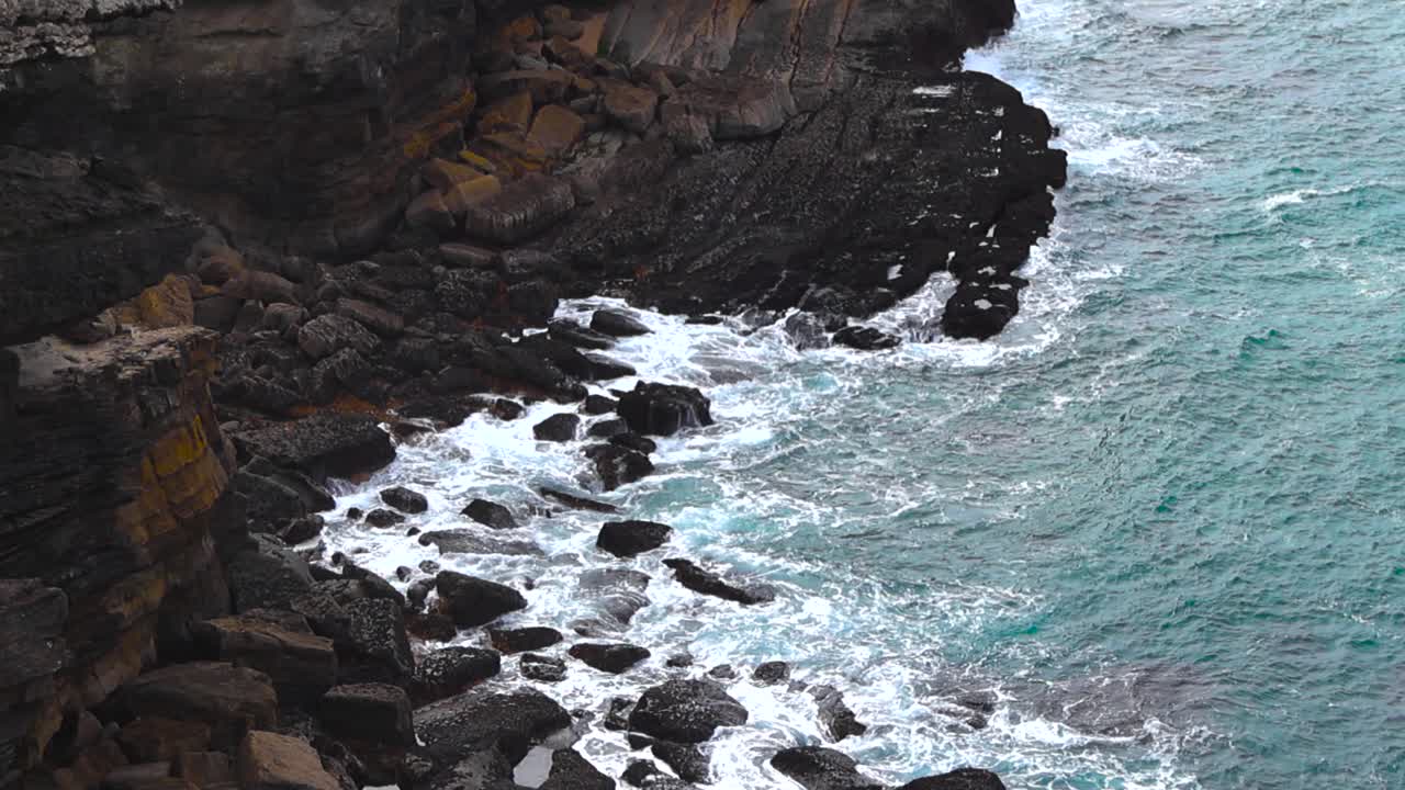 Ripresa dall'alto con movimento panoramico lento di onde turchesi dell'Atlantico che si infrangono contro le aspre scogliere rocciose lungo la pittoresca costa di Azenhas do Mar in Portogallo
