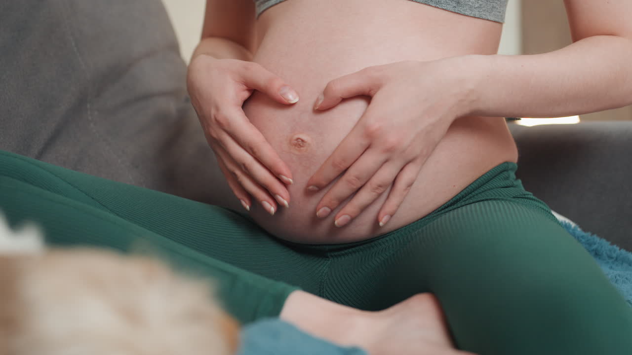 Donna incinta seduta sul divano che massaggia la pancia con respirazione lenta, rituali calmanti, top sportivo e leggings verdi, pelle morbida, soggiorno accogliente, pratica di rilassamento prenatale, connessione di accudimento.