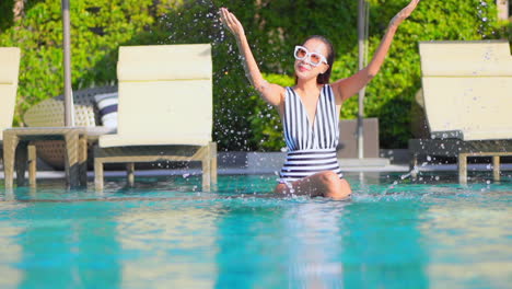 playful attractive asian woman playing water splash smiling sitting at shallow water of swimming pool next to deckchairs at hotel lounge, slow-motion
