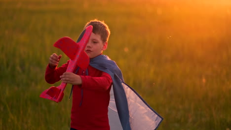 Der-Junge-Pilot-Läuft-In-Einem-Roten-Regenmantel-Mit-Einem-Flugzeug-In-Der-Hand-Und-Lacht-Bei-Sonnenuntergang-Auf-Der-Sommerwiese.-Er-Stellt-Sich-Vor,-Er-Sei-Ein-Flugzeugpilot,-Der-Mit-Einem-Modellflugzeug-Spielt