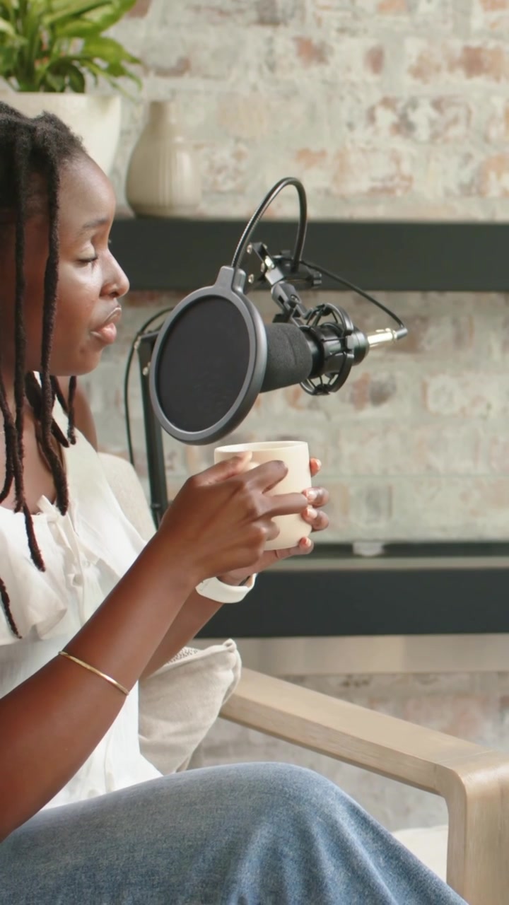Video verticale: Parla a comando, conduttore con dreadlocks in maglietta bianca che fa un podcast al microfono, sedia, tazza, orologio