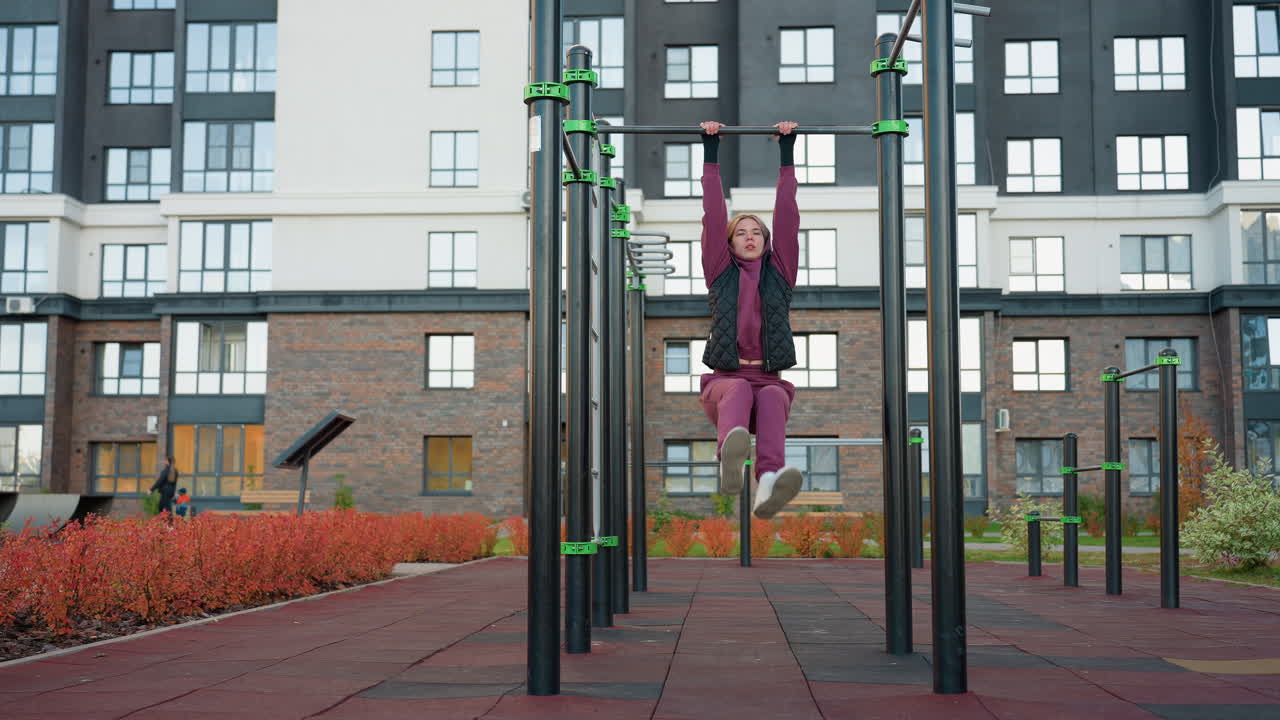 Outdoor urban park workout scene featuring female athlete in hoodie and vest hanging from pull up bar and lifting legs to engage core muscles and build strength, endurance and balance