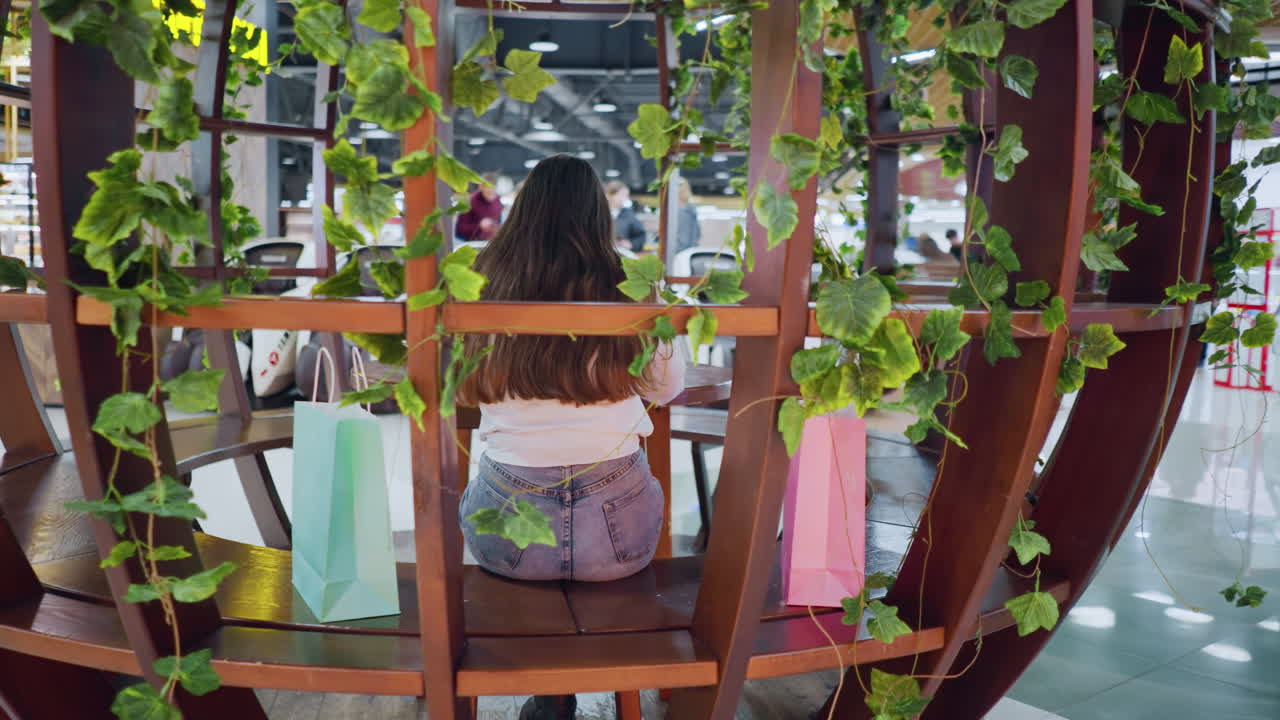 고급스러운 식당에서 vip 좌석에 앉아있는 여자의 뒷면, 그녀의 에 쇼핑 가방과 함께, 그녀의 전화에 초점을 맞추고, 그녀를 둘러싼 사람들, 디지털 불빛이 배경에서 박이고