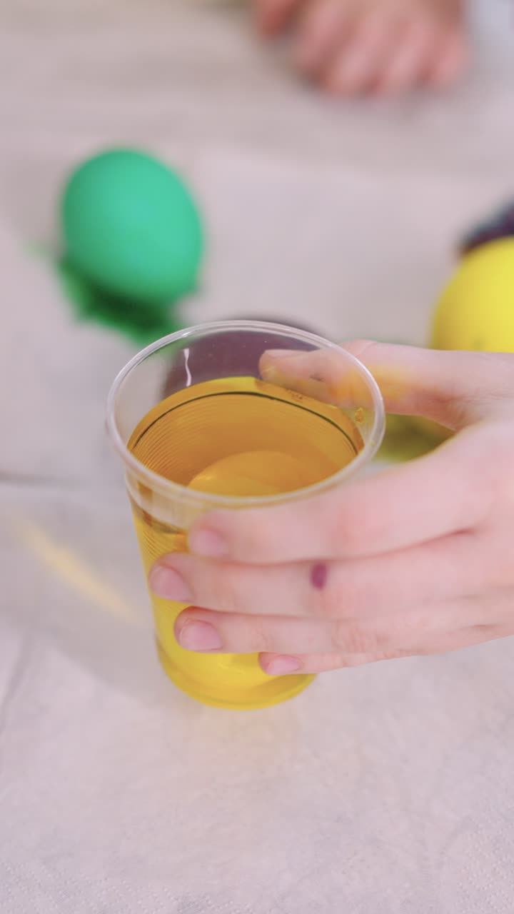 Manos sumergiendo un huevo de Pascua en tinte amarillo en una taza.