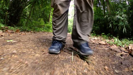 un hombre caminando en la jungla, la cámara enfocada en sus piernas, mostrándolo dando un paso adelante en el camino de tierra con sus zapatos enmarcados
