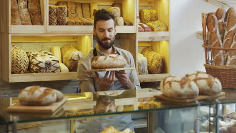 Gut-Aussehender-Junger-Männlicher-Bäckereiverkäufer,-Der-Morgens-Frisches-Brot-An-Der-Theke-Riecht-Und-Vor-Der-Kamera-Posiert