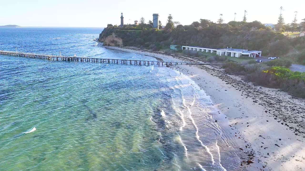 Drone footage captures a serene beach and pier on Bellarine Peninsula, Victoria, with clear waters and lush greenery under soft daylight