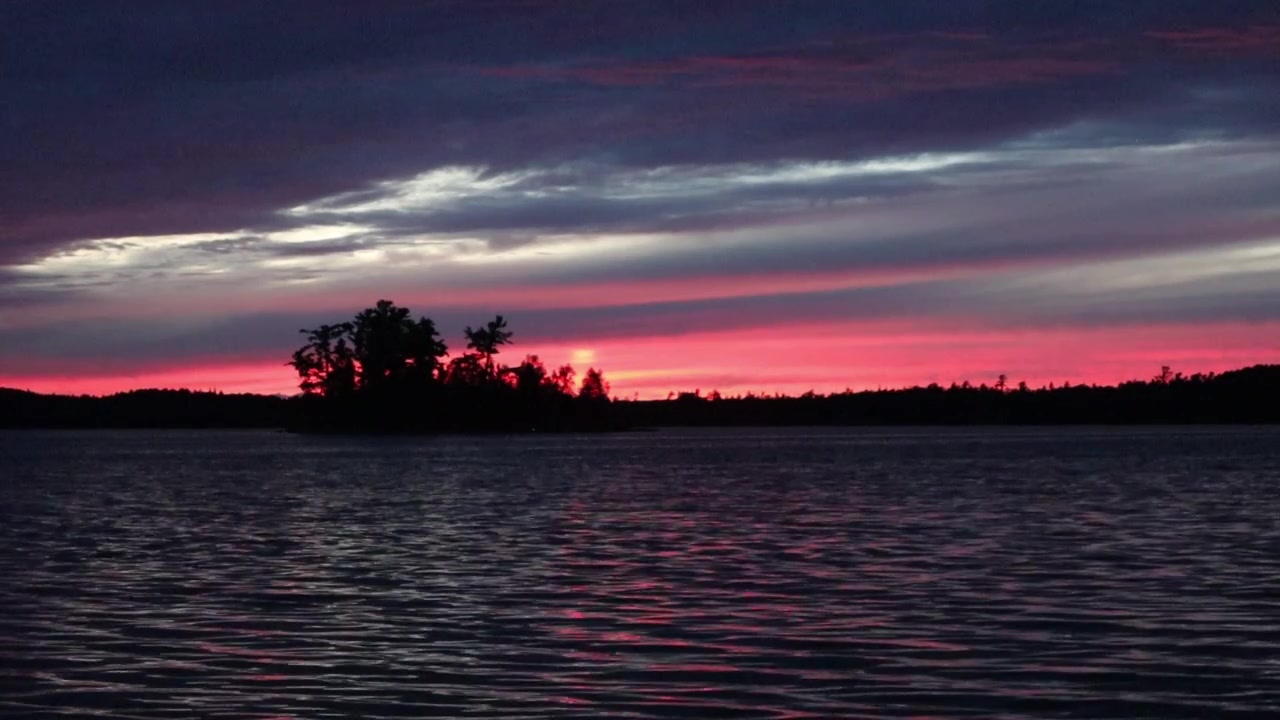Sunset at Caliper Lake in Northwest Ontario, Canada. Red, orange, and blue colors fill the sky as waves move across the lake with trees silhouette along the distant shoreline.