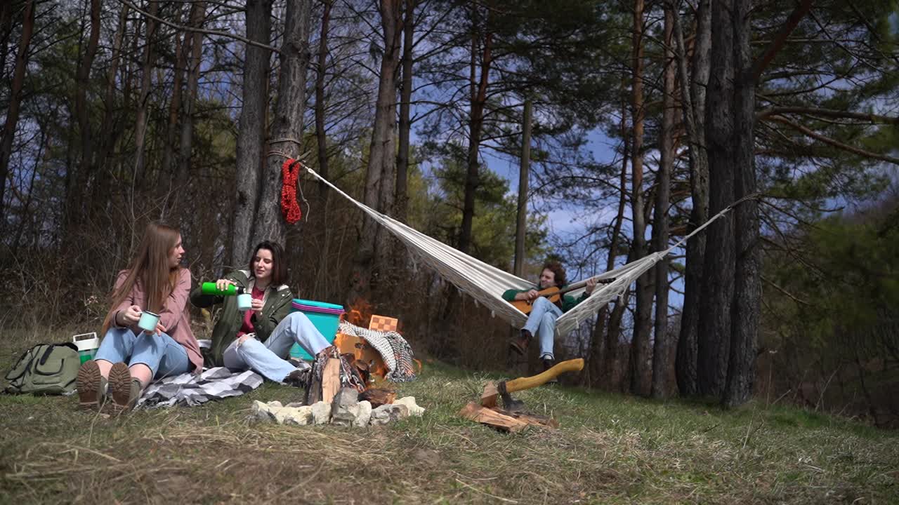 две девушки и юноша весело проводят время в лесу. девушки пьют горячий напиток, а юноша играет на гитаре.