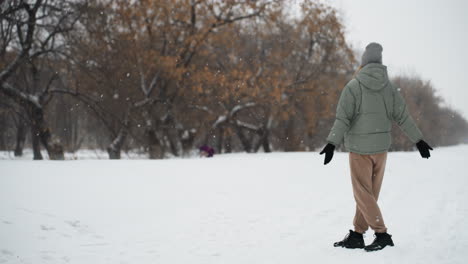 woman in green winter coat and gray beanie stands with arms outstretched in snowy park while snow falls gently, dog in purple coat visible in distance near forest edge, serene cold weather scene