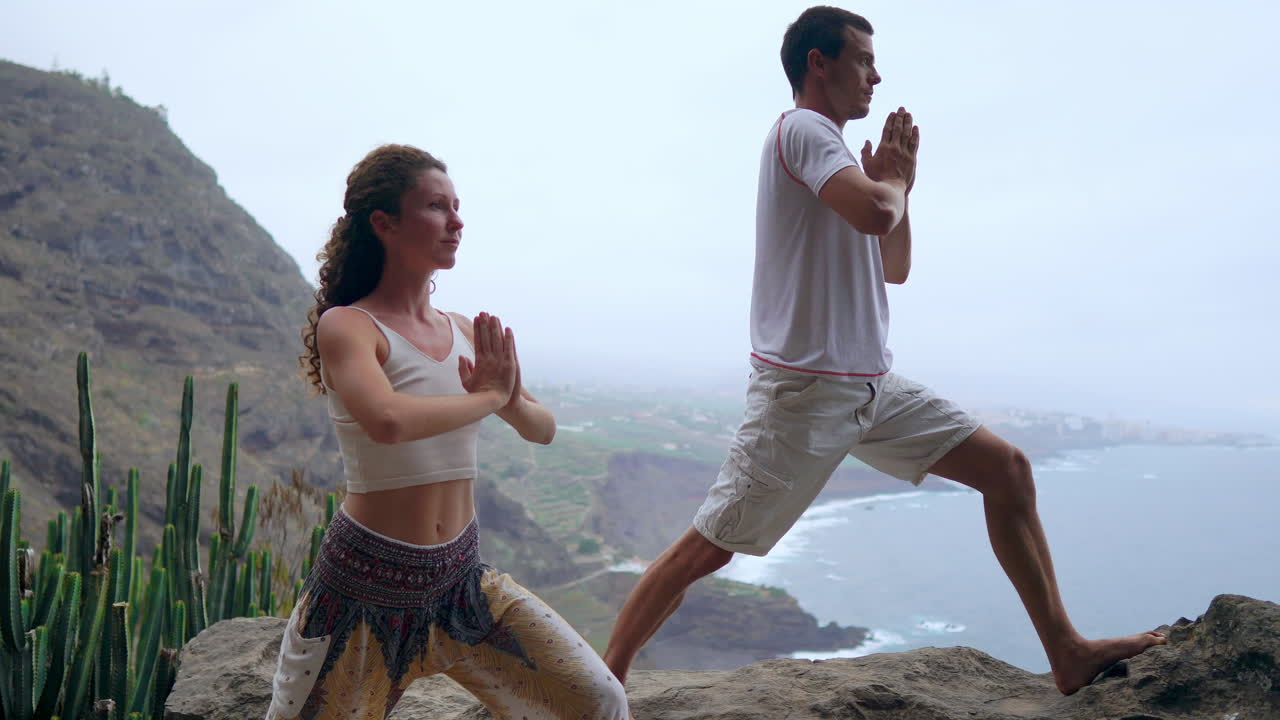 Positioned on a cliff's brink, a man and woman inhale the sea air, their hands raised during a yoga session with the ocean view before them